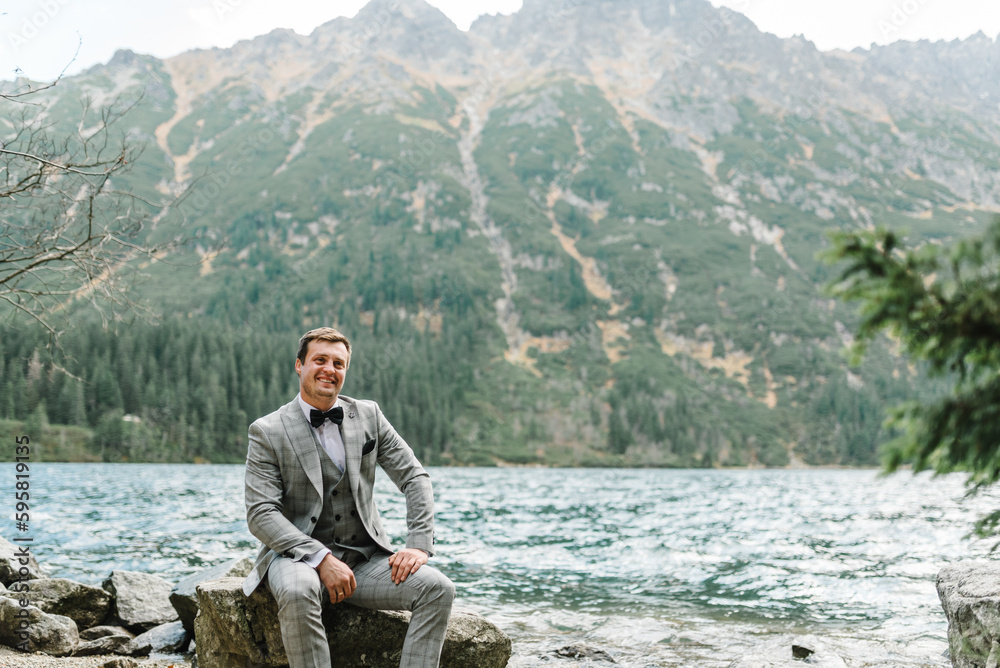 Fototapeta premium Man on the stony shore. The groom near the lake in the mountains. Groom against the backdrop of a mountain landscape.