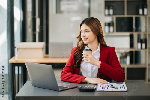 Confident beautiful Asian businesswoman typing laptop computer and digital tablet while holding coffee at modern office