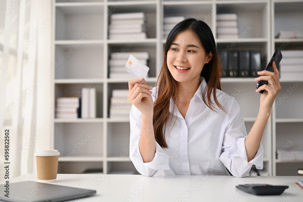 Beautiful young Asian female office employee holding her credit card and smartphone Beautiful young Asian female office employee holding her credit card and smartphone