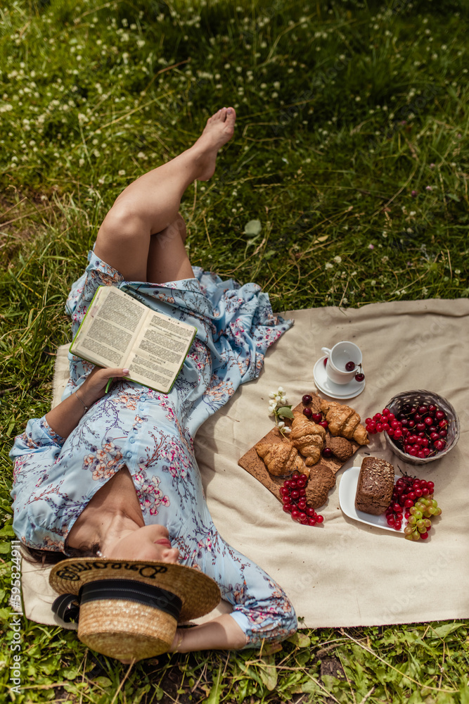 Girl in straw hat reads book lying on blanket in nature in summer at picnic