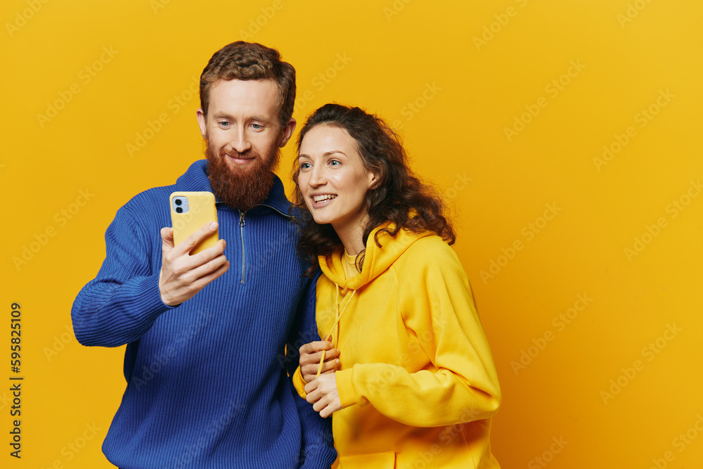 Man and woman couple smiling merrily with phone in hand social media viewing photos and videos, on yellow background, symbols signs and hand gestures, family freelancers. Man and woman couple smiling merrily with phone in hand social media viewing photos and videos, on yellow background, symbols signs and hand gestures, family freelancers.
