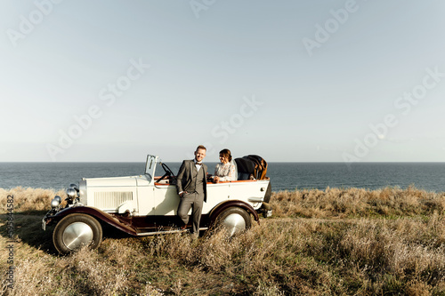 an amazing wedding couple on top of a hill overlooking the sea stands near the car. bride and groom at sunset near the sea
