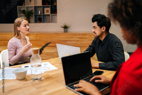Remote view of elegant male team leader sitting at table with multiethnic colleagues, discussing working issues at negotiations meeting. Serious male businessman talking at diverse group meeting.