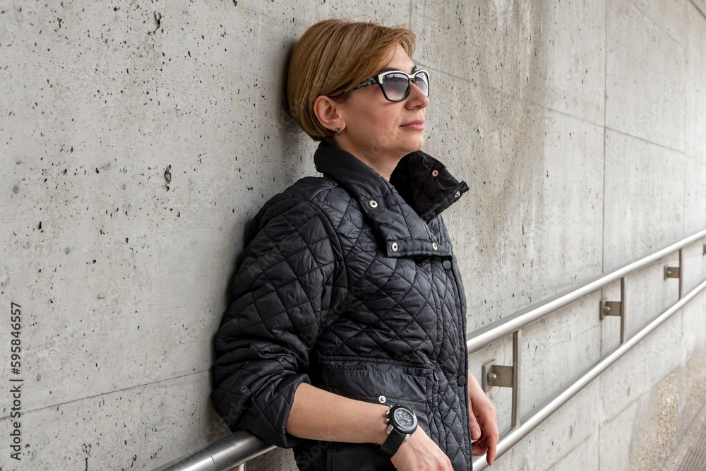 Street portrait of a businesswoman 35-40 years old at a concrete wall in a black jacket and glasses.