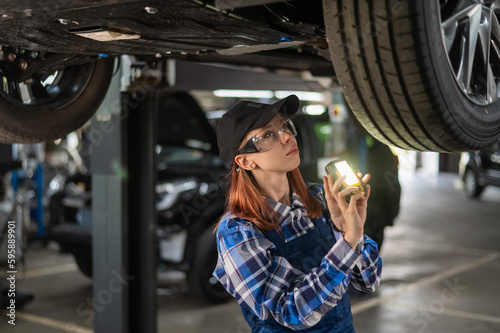 A female mechanic inspects a lifted car. A girl at a man's work. © Михаил Решетников