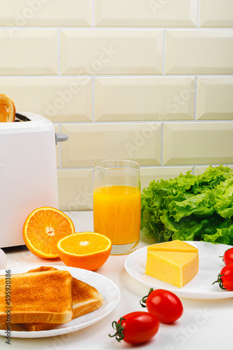 Healthy breakfast in the kitchen. Toaster and bread toast, eggs, tomatoes, freshly squeezed orange juice. Cheese, butter, lettuce. White toaster with healthy food and drinks on table in light kitchen