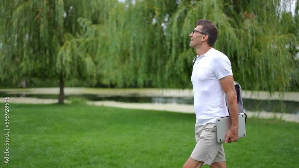 Side view happy man with laptop and backpack walking on the right at background of spring summer landscape leaving in slow motion. tracking shot confident successful Caucasian freelancer outdoors Side view happy man with laptop and backpack walking on the right at background of spring summer landscape leaving in slow motion. tracking shot confident successful Caucasian freelancer outdoors
