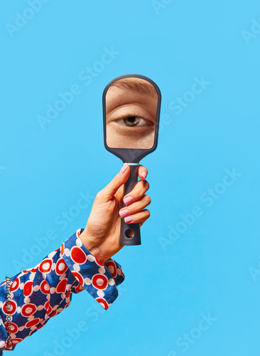 Female hand holding small mirror with reflection of girl's eye without makeup over bright blue background. Vintage style photo