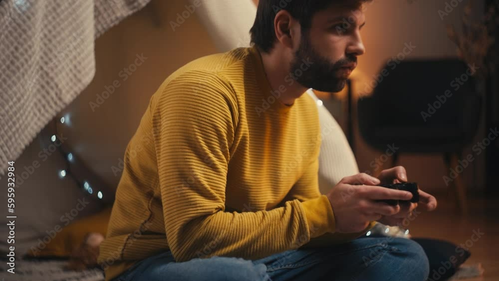 Young man playing video game sitting inside blanket fort, reliving childhood Young man playing video game sitting inside blanket fort, reliving childhood