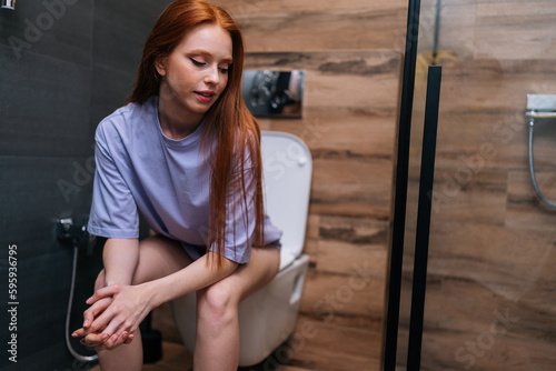 Positive young woman in casual clothes sitting on toilet bowl at home bathroom with modern interior, smiling looking away. Tired exhausted redhead female sitting in restroom after party feeling sleepy