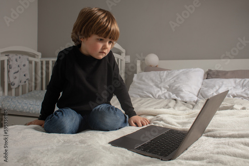 Little boy sitting on bed and looking at laptop. The kid watches cartoons and plays on the computer. Toddler learns something new and interesting.