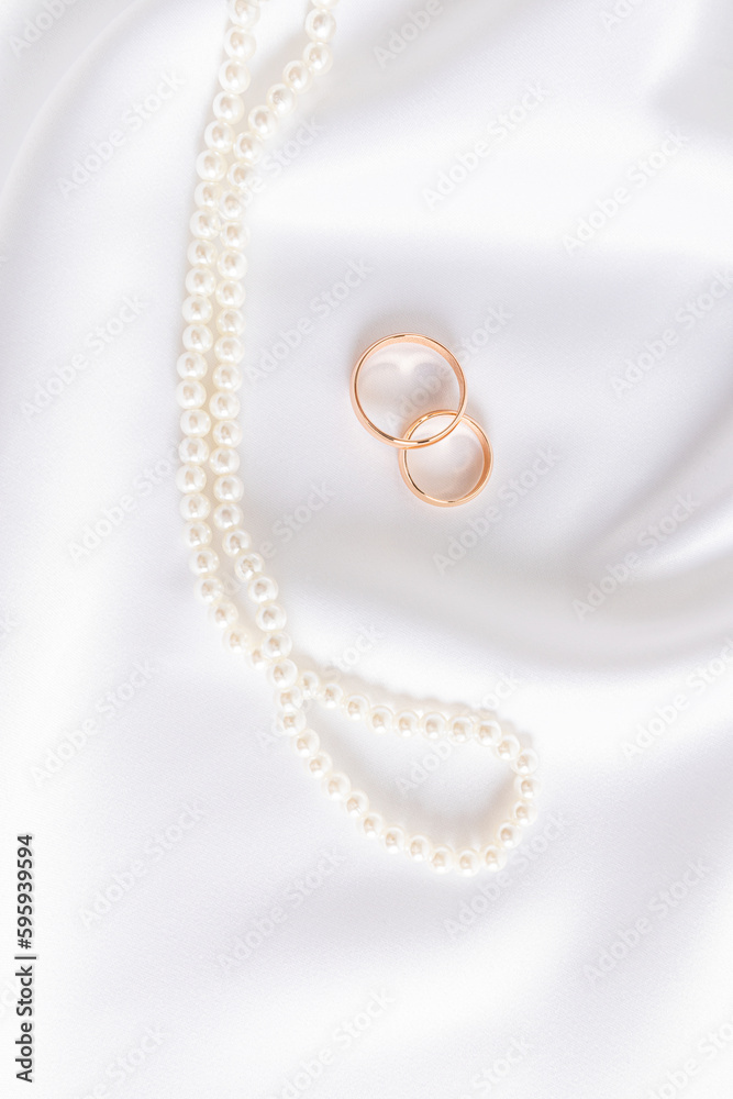 Two gold wedding rings on a chic white satin background with a pearl necklace. Vertical wedding background. layout for design. Two gold wedding rings on a chic white satin background with a pearl necklace. Vertical wedding background. layout for design.