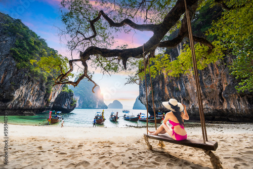 Young woman in bikini sitting on a swing against white beach and blue sea at Koh Lao Lading, Krabi, Thailand.