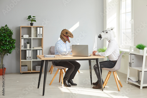 Two funny business men wearing animal masks sitting at the desk of their workplace with laptop and having discussion in office, considering new projects or startups, analyzing company.