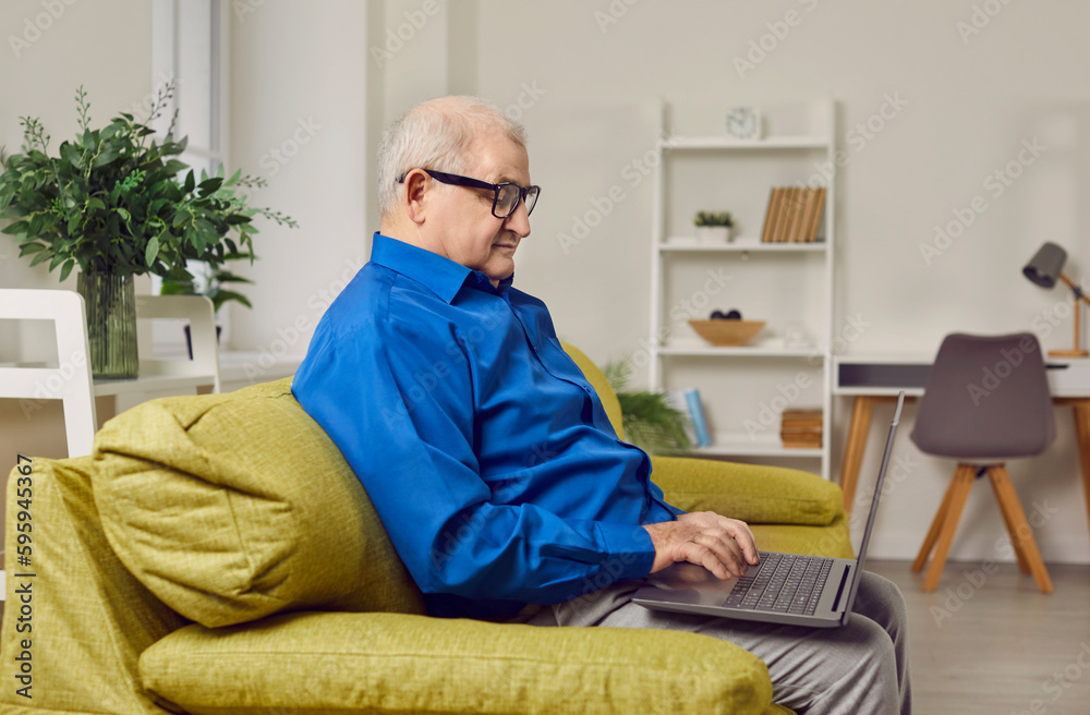 Focused elderly man sitting on sofa using laptop. Side view portrait of senior man with eyeglasses wearing blue shirt holding laptop on his laps and typing while sitting on couch at home