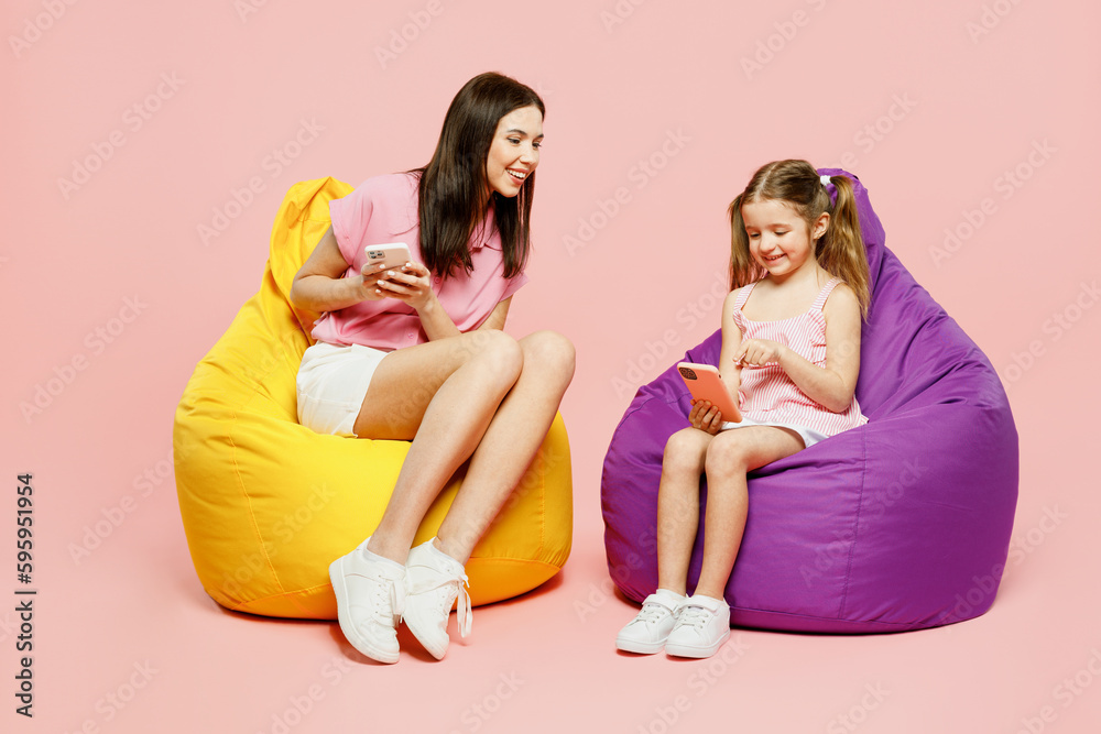 Full body happy woman wear casual clothes with child kid girl 6-7 years old. Mother daughter sit in bag chair hold use mobile cell phone isolated on plain pink background. Family parent day concept. - obrazy, fototapety, plakaty 