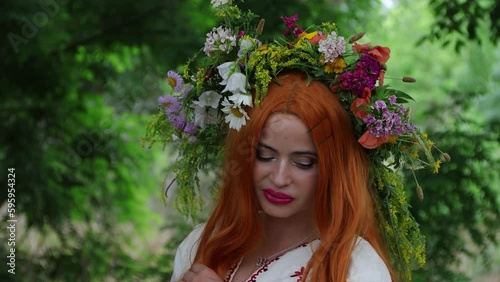 Beautiful girl with long red hair with flower crown in embroidered ethnic suit stands by tree. Fairytale character, forest spirit, mavka, fairy. 4k resolution