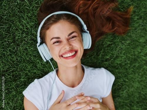 Beautiful woman in headphones listening to music lying on the grass green lawn in the park in the summer and laughing, happiness in nature