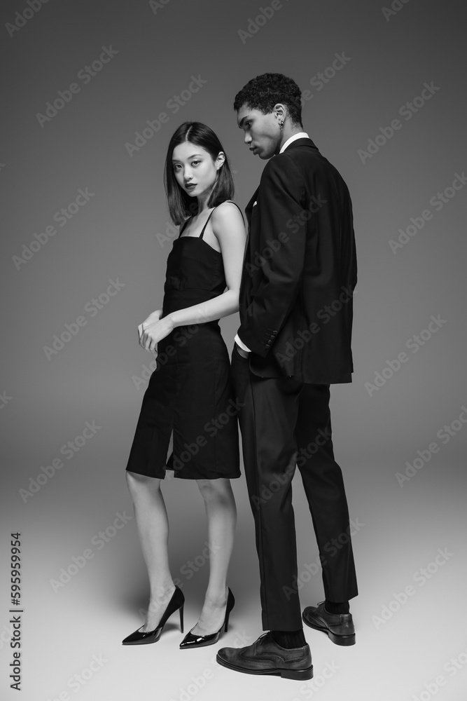 full length of asian woman in black elegant dress looking at camera near african american man in suit on grey background. full length of asian woman in black elegant dress looking at camera near african american man in suit on grey background.