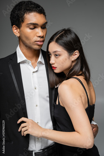 elegant african american guy in black blazer embracing sensual asian woman looking away isolated on grey.