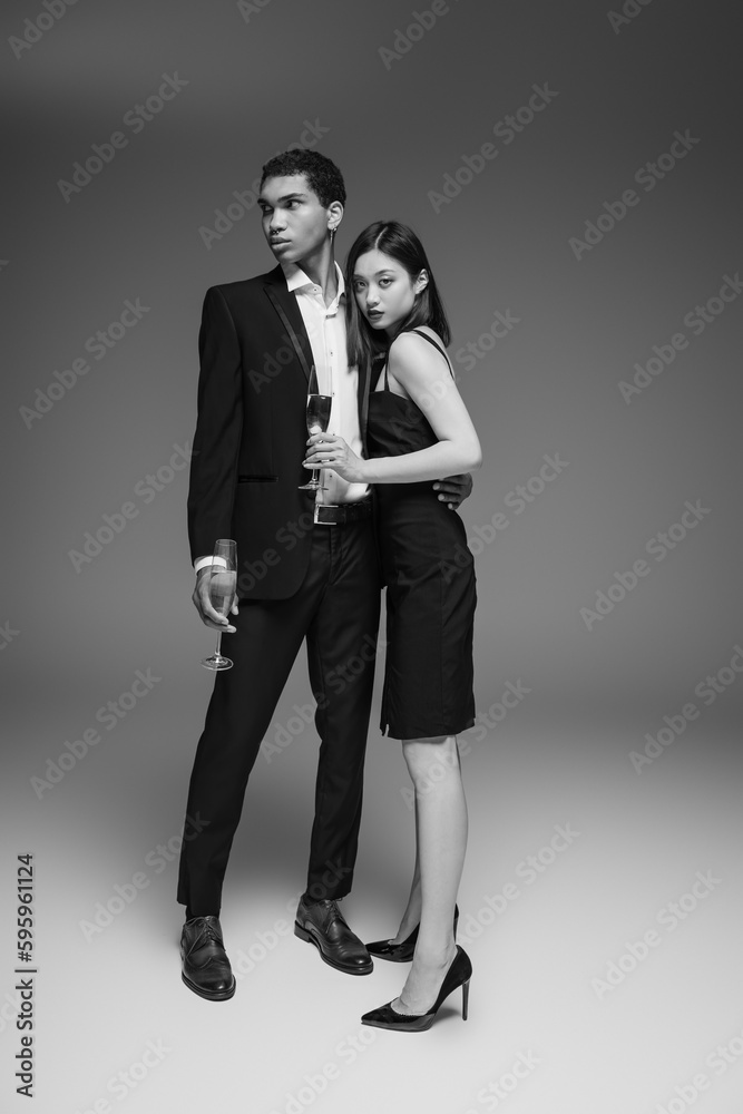 full length of interracial couple in elegant and stylish attire standing with champagne glasses on grey background.