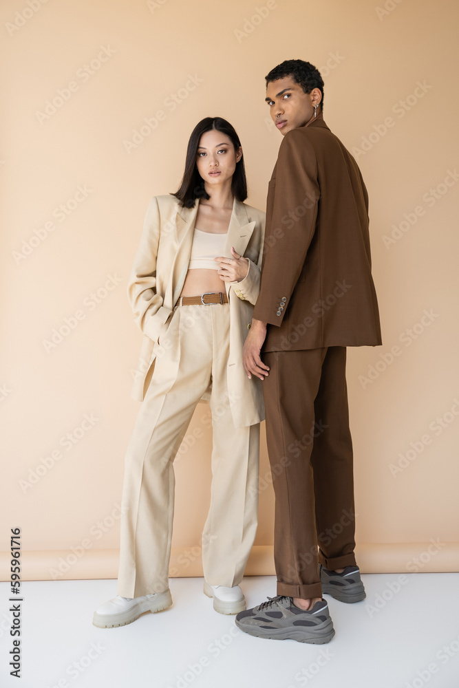© LIGHTFIELD STUDIOS - full length of young interracial couple in trendy pantsuits looking at camera while posing on beige background. © LIGHTFIELD STUDIOS - full length of young interracial couple in trendy pantsuits looking at camera while posing on beige background.