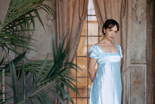 A young beautiful girl in a blue Regency dress stands at a wooden window in the estate. The classic artistic image of Russian literature of the 19th century, outdated.