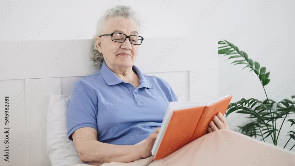 An elderly woman with glasses sits in bed in her bedroom, reading a book with interest and a smile An elderly woman with glasses sits in bed in her bedroom, reading a book with interest and a smile