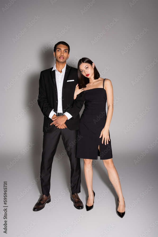 full length of interracial couple in elegant formal wear looking at camera on grey background. full length of interracial couple in elegant formal wear looking at camera on grey background.