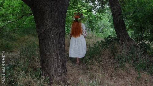 Paganism. Beautiful girl with long red hair with flower crown in embroidered ethnic suit walks in forest among trees. Fairytale character, forest spirit, mavka, fairy. 4K Resolution