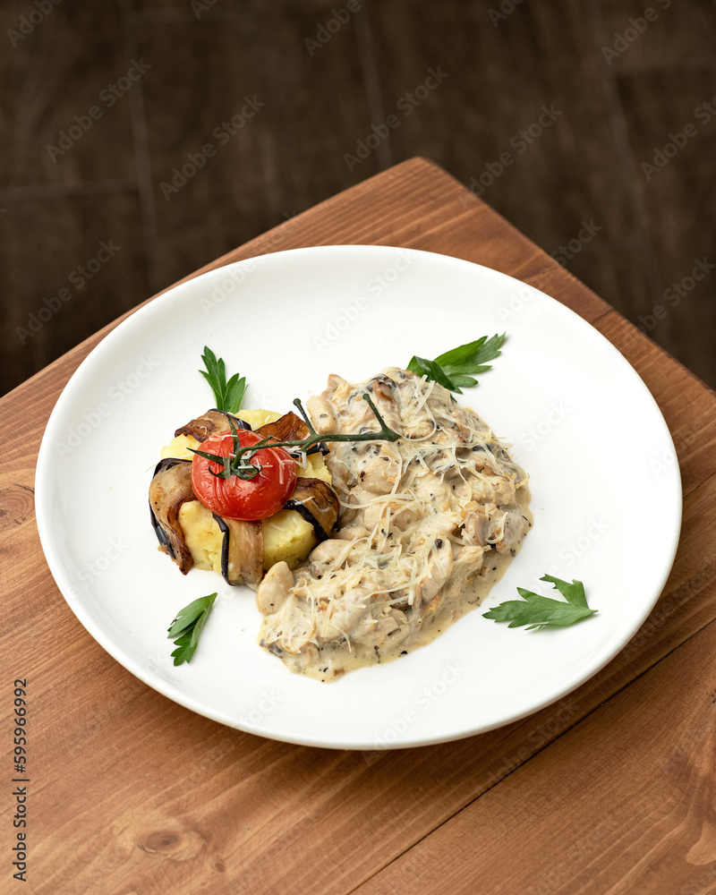 Mushrooms in creamy sauce with cheese and garnish of mashed potatoes, eggplant and tomato. Second course on white plate. Vegetarian dish. Wooden background. View from above. Mushrooms in creamy sauce with cheese and garnish of mashed potatoes, eggplant and tomato. Second course on white plate. Vegetarian dish. Wooden background. View from above.
