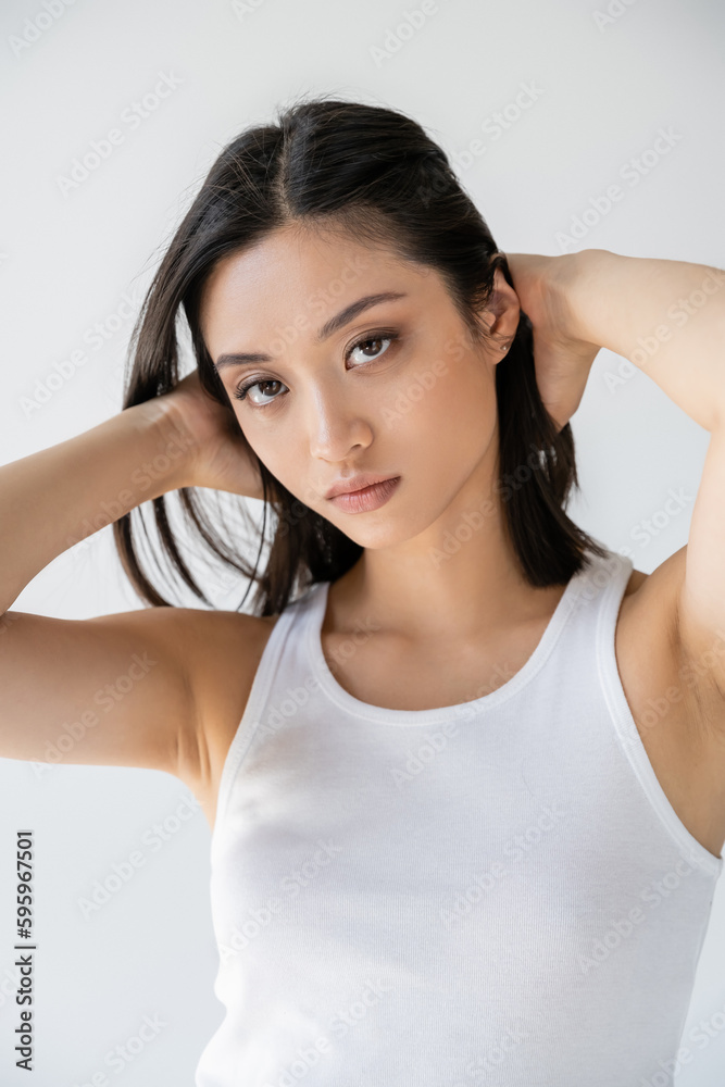 young asian woman in white tank top touching brunette hair and looking at camera isolated on grey. young asian woman in white tank top touching brunette hair and looking at camera isolated on grey.