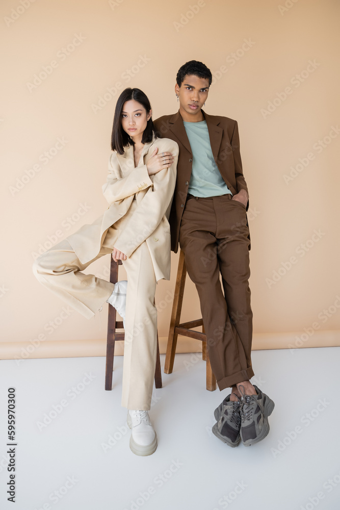 full length of young multiethnic couple in stylish suits posing near stools on beige background. full length of young multiethnic couple in stylish suits posing near stools on beige background.