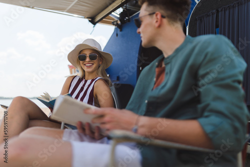 Young couple sitting together in front of van, camping and reading book.
