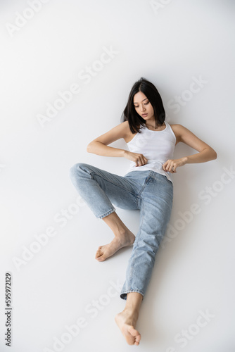 full length of barefoot asian woman in white tank top and jeans lying on grey background.