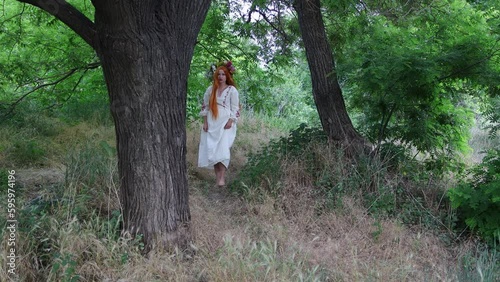 Paganism. Beautiful girl with long red hair with flower crown in embroidered ethnic suit walks in forest among trees. Fairytale character, forest spirit, mavka, fairy. 4K Resolution