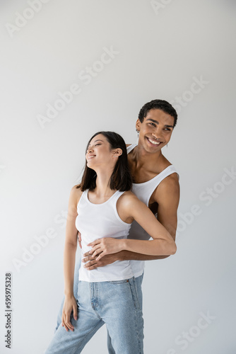happy african american guy hugging asian woman smiling with closed eyes isolated on grey.