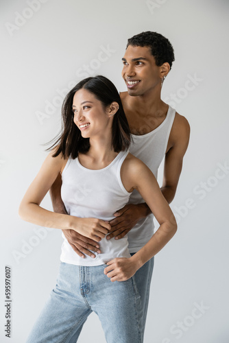 young african american man embracing carefree asian model in jeans and white tank top isolated on grey.