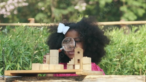 Young black girl is using a magnifying glass to perform analytical gestures playing with wooden blocks.
