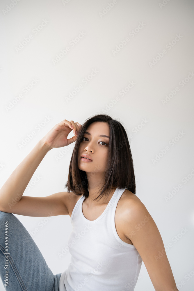 brunette asian woman in white tank top holding hand near face and looking at camera on grey background. brunette asian woman in white tank top holding hand near face and looking at camera on grey background.