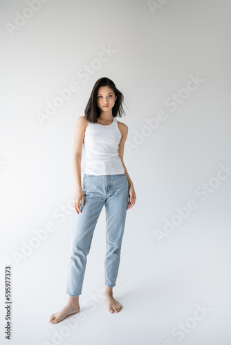 full length of barefoot and slim asian woman in tank top and jeans standing on grey background.