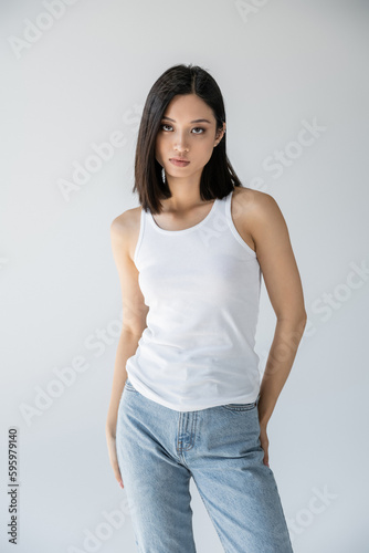 sensual asian woman in jeans and white tank top looking at camera isolated on grey.