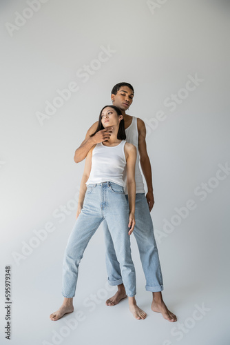 full length of stylish african american man touching chin of barefoot asian woman in tank top and jeans on grey background.