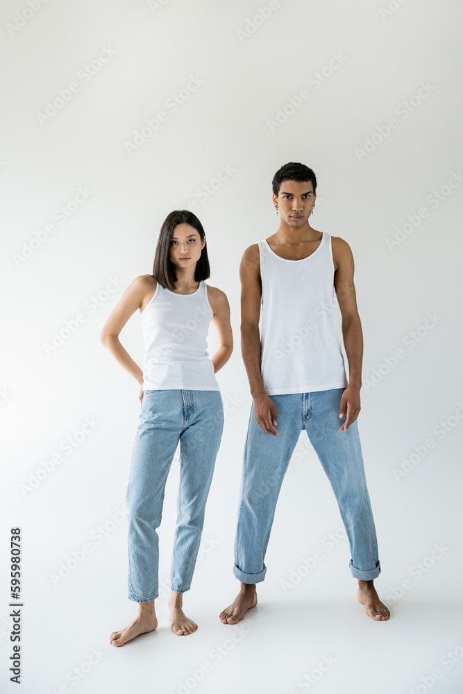 full length of young and barefoot multiethnic couple posing in white tank tops and blue jeans on grey background. full length of young and barefoot multiethnic couple posing in white tank tops and blue jeans on grey background.