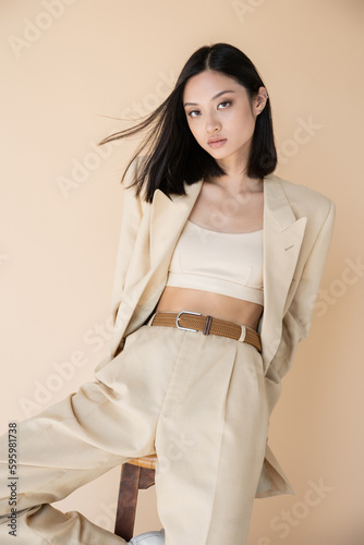 Foto fashionable asian woman in pantsuit posing near stool and looking at camera isolated on beige