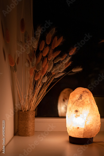 Warm light from a salt lamp illuminating a bouquet of lagurus in the evening. Warm light. Room lighting. Home comfort. The benefits of a salt lamp. Dry flowers in the interior. Apartment decoration.