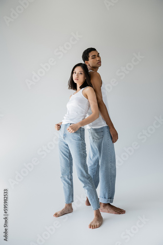 full length of barefoot interracial couple in white tank tops and jeans posing back to back on grey background.