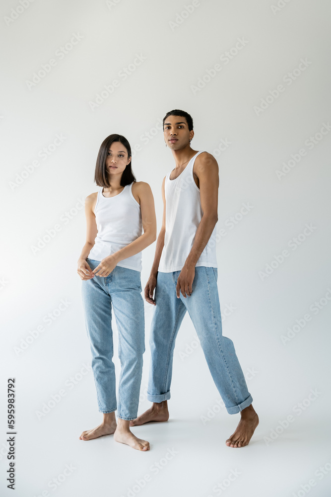 full length of barefoot and slender interracial couple in white tank tops and jeans posing on grey background. full length of barefoot and slender interracial couple in white tank tops and jeans posing on grey background.