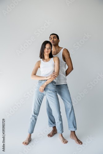 full length of barefoot and slender multiethnic couple in tank tops and jeans on grey background.
