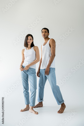 full length of barefoot and slender interracial couple in white tank tops and jeans posing on grey background.